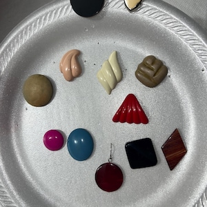 May include: A collection of colorful, vintage-style earrings and buttons. The assortment includes a black circular button, a red triangular earring, and a brown diamond-shaped earring. Other shapes include a pink circle, a blue oval, and a cream-colored geometric shape.