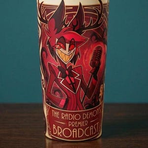 May include: A cylindrical tumbler featuring a graphic illustration of a red-haired character with antlers holding a vintage microphone. The design includes the text "THE RADIO DEMON'S PREMIER BROADCAST" in a decorative font against a deep red background.