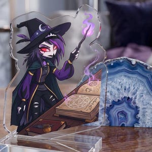 May include: A clear acrylic standee featuring a cartoon character dressed as a witch with purple hair, a black hat, and a dark cloak. The character holds a wand emitting purple magic over an open spellbook with a circular pentagram symbol.