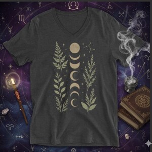 May include: Dark gray V-neck t-shirt featuring a design of moon phases and botanical elements in beige and light green. The shirt has a celestial theme, with a background of astrological symbols.