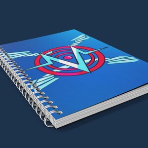 May include: A spiral-bound notebook with a blue cover featuring a graphic design of a red and blue circle with a lightning bolt and wings. The notebook is on a dark blue surface.