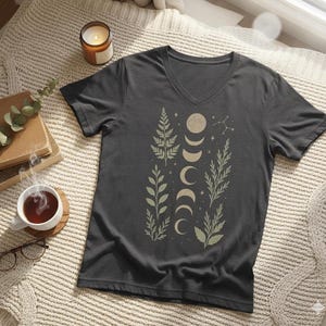 May include: Dark gray V-neck t-shirt featuring a celestial design with moon phases and botanical elements in a muted green color. The design includes crescent moons, stars, and various leaf and fern illustrations. The shirt is made of a soft material.