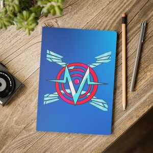 May include: A blue notebook with a graphic design featuring a red circle, a teal and white V-shaped design, and teal wing-like shapes. A pencil and pen are next to the notebook on a wooden surface.