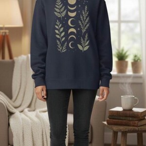 May include: Navy blue sweatshirt with a gold moon phase and botanical design. The long-sleeved top features a crew neck and a relaxed fit. The design includes crescent moons, stars, and leafy branches. The model is wearing black jeans and white sneakers.