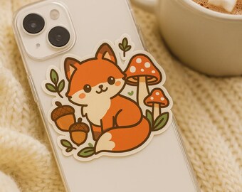 Op de afbeelding: Een doorzichtig telefoonhoesje met een sticker met een cartoonvos, paddenstoelen en eikels. De vos is oranje en wit, met een vriendelijke uitdrukking. De paddenstoelen hebben rode en witte stippen. Op de achtergrond staat een mok warme chocolademelk met marshmallows.
