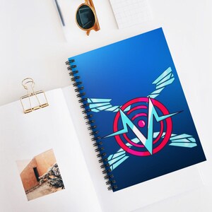 May include: Spiral-bound notebook with a vibrant blue cover featuring a geometric design. The design includes a red and pink circle with a light blue 'N' shape and angular accents. The notebook is open on a white surface.