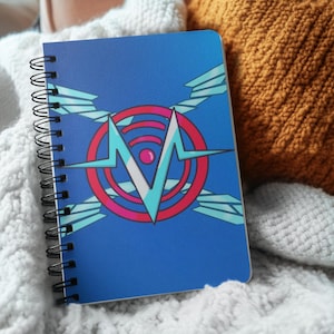 May include: A spiral-bound notebook with a blue cover featuring a graphic design. The design includes a red circle with a white and teal emblem, and teal accents. The notebook is resting on a white knitted blanket.