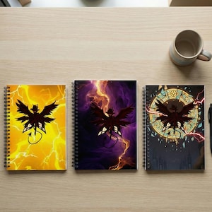 May include: Three spiral-bound notebooks with a dark silhouette of a winged creature. The covers feature vibrant backgrounds of yellow lightning, purple clouds, and a stained-glass design. A pen and mug are also on the wooden surface.