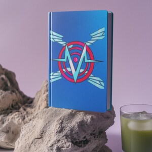 May include: A blue notebook with a geometric design featuring a red circle, a white "M", and light blue wings. The notebook is resting on a light-colored rock, with a glass of green liquid with ice in the background.