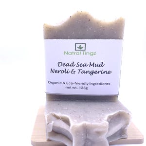 May include: A light grey bar of soap, labelled "Dead Sea Mud Neroli & Tangerine" by Natral Tingz. The label also states "Organic & Eco-friendly Ingredients" and "net wt. 125g". The soap is on a small wooden surface.