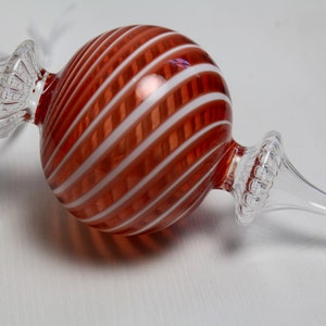 以下が含まれることがあります： 赤と白のストライプ模様の透明なガラス製のクリスマスツリーオーナメント。オーナメントは丸い形で、透明なガラスの茎と装飾的なアクセントが付いています。オーナメントの長さは約15cmです。