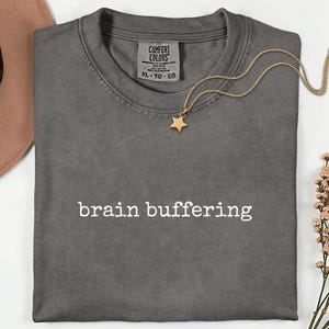 May include: A grey Comfort Colors t-shirt with the words "brain buffering" printed in white. A gold-coloured star pendant necklace rests on the shirt. A brown hat and floral arrangement are in the background.