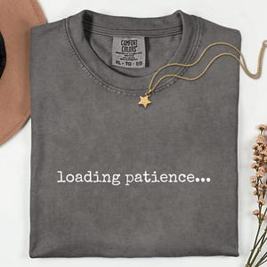 May include: A grey Comfort Colors t-shirt with the words "loading patience..." in white. A gold star pendant necklace is on the shirt. A brown hat and a sprig of flowers are in the background.