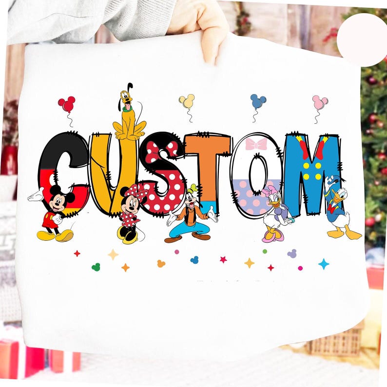 May include: A white sweatshirt featuring the word CUSTOM in large, colorful, patterned letters. Mickey Mouse, Minnie Mouse, Pluto, Goofy, Daisy Duck, and Donald Duck are positioned around the letters, accompanied by small floating balloons.