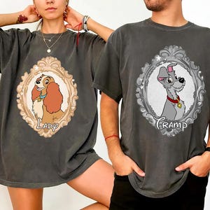 K&ouml;nnte beinhalten: Zwei anthrazitfarbene T-Shirts mit Cartoon-Portr&auml;ts von Hunden in verzierten Rahmen. Eines zeigt "Lady" und das andere "Tramp" in Schrift. Die Shirts haben kurze &Auml;rmel und eine lockere Passform.