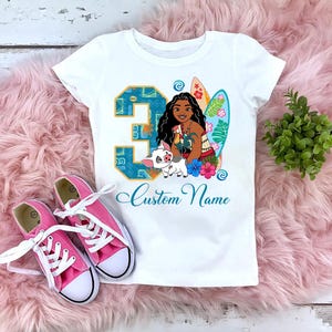 Puede incluir: Camiseta blanca con un gr&aacute;fico colorido de un personaje de dibujos animados, un cerdo y una tabla de surf. El n&uacute;mero "3" est&aacute; en un dise&ntilde;o azul. El texto "Custom Name" est&aacute; escrito en cursiva. Zapatillas de lona rosas con cordones blancos junto a la camiseta.