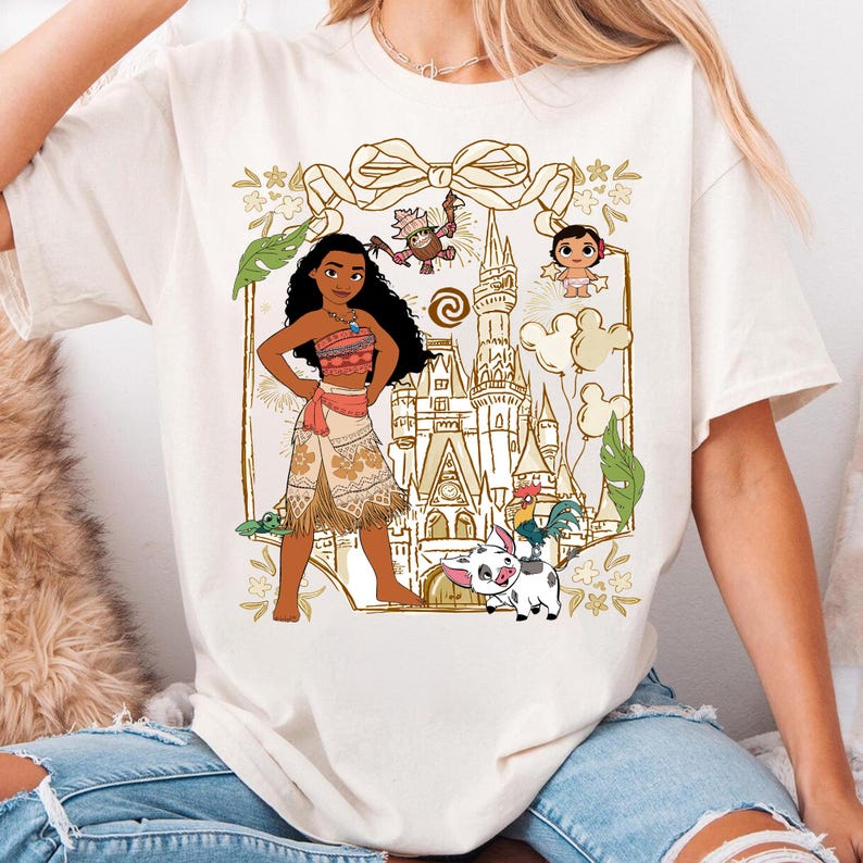 May include: Cream-colored t-shirt featuring a graphic of a woman in a traditional outfit, a castle, and cartoon characters. The design includes a bow, flowers, and other decorative elements. The shirt is made of a soft material.