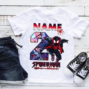 Może przedstawiać: Biała koszulka z grafiką Spider-Mana, cyfrą 2 i napisem "NAME" w kolorze czerwonym i czarnym. Zestaw zawiera ciemnoniebieskie dżinsowe szorty i czarne pł&oacute;cienne trampki z białymi podeszwami i sznurowadłami.