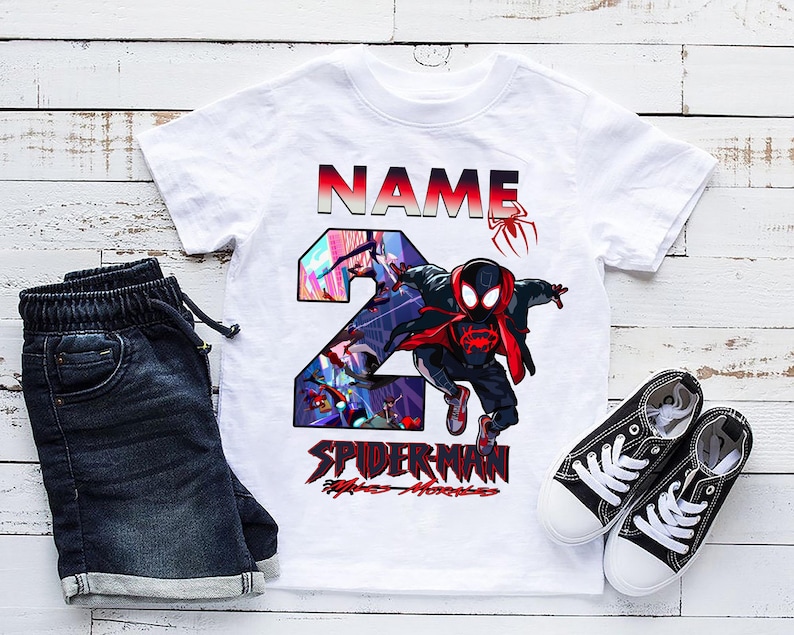 Może przedstawiać: Biała koszulka z grafiką Spider-Mana, cyfrą 2 i napisem "NAME" w kolorze czerwonym i czarnym. Zestaw zawiera ciemnoniebieskie dżinsowe szorty i czarne pł&oacute;cienne trampki z białymi podeszwami i sznurowadłami.