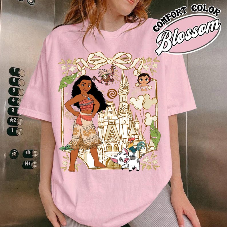 May include: Pink t-shirt featuring a graphic of a cartoon character with a castle, characters, and the text "COMFORT COLOR Blossom". The design includes a bow and floral elements. The shirt is a light pink color.