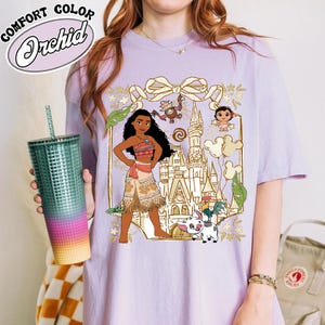 May include: A light purple t-shirt featuring a graphic of a cartoon character with a castle, animals, and floral accents. The shirt has the words "Comfort Color Orchid" in a white, cursive font. A teal tumbler with a straw is held in the image.