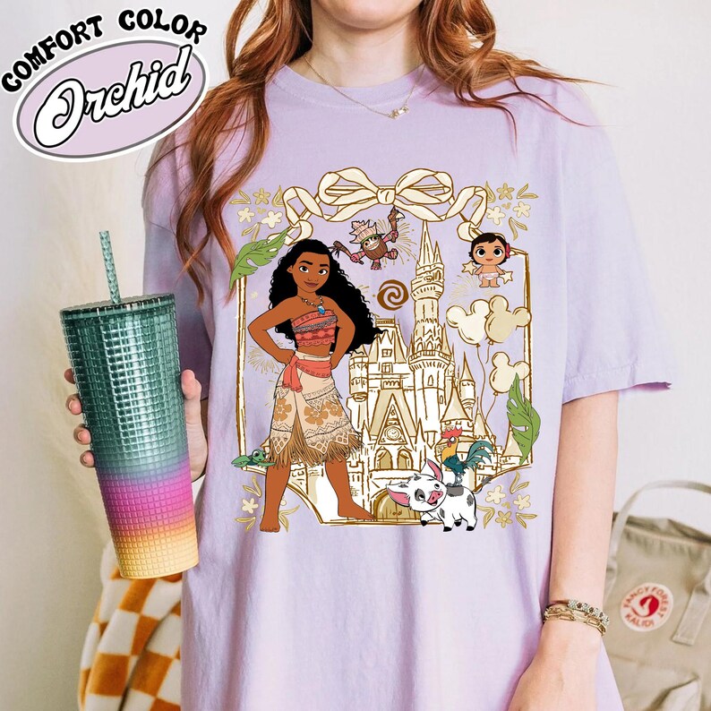 May include: A light purple t-shirt featuring a graphic of a cartoon character with a castle, animals, and floral accents. The shirt has the words "Comfort Color Orchid" in a white, cursive font. A teal tumbler with a straw is held in the image.