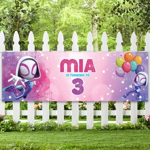 Pode incluir: Banner de anivers&aacute;rio com um fundo gradiente rosa e roxo. Exibe o nome "MIA" e o texto "IS TURNING TO 3". O banner apresenta ilustra&ccedil;&otilde;es de personagens de super-her&oacute;is e bal&otilde;es, ideal para uma festa de anivers&aacute;rio.