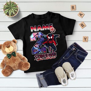 Może przedstawiać: Czarny t-shirt dla malucha z grafiką Spider-Mana, cyfrą 2 i sekcją na imię. Koszulka jest połączona z niebieskimi dżinsami i beżowymi butami. Na zdjęciu znajduje się r&oacute;wnież pluszowy miś i roślina doniczkowa.