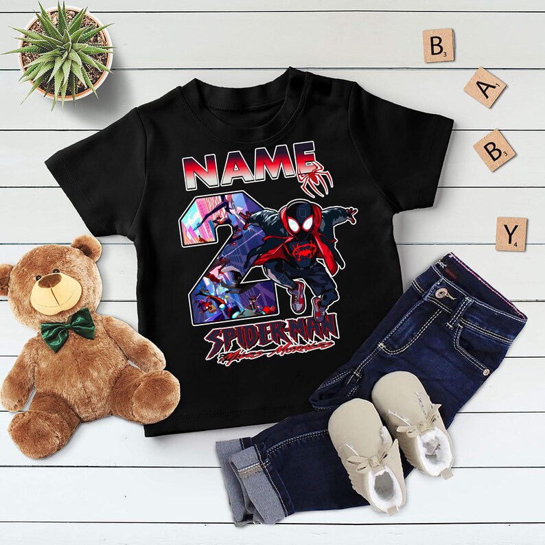 Może przedstawiać: Czarny t-shirt dla malucha z grafiką Spider-Mana, cyfrą 2 i sekcją na imię. Koszulka jest połączona z niebieskimi dżinsami i beżowymi butami. Na zdjęciu znajduje się r&oacute;wnież pluszowy miś i roślina doniczkowa.