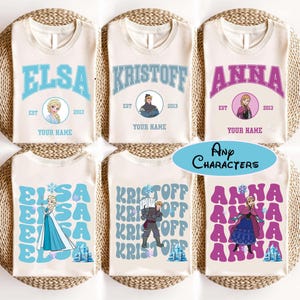 Puede incluir: Camisetas color crema con los nombres "ELSA", "KRISTOFF" y "ANNA" en varias fuentes y colores. Cada camiseta incluye una imagen de personaje, el a&ntilde;o "2013" y el texto "YOUR NAME". Las camisetas se muestran sobre una superficie tejida.