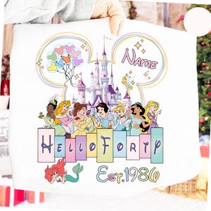 May include: White sweatshirt featuring a colorful Disney castle, princess illustrations, and the text "Hello Forty Est. 1986". Includes a customizable name field and Mickey Mouse ears design.