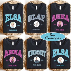 Puede incluir: Camisetas negras con nombres de personajes en texto arqueado. Cada camiseta presenta un nombre diferente: Elsa, Olaf, Anna y Kristoff. Cada dise&ntilde;o incluye una imagen del personaje, el a&ntilde;o 2013 y el texto "Your Name". Tambi&eacute;n est&aacute; presente el texto "Any Characters".