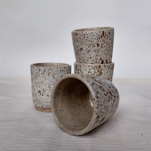 May include: Four speckled ceramic cups in neutral tones. The cups have a cylindrical shape with a slight taper and a speckled pattern in brown and beige. One cup is on its side, the others are upright. Each cup is approximately 8 cm tall.