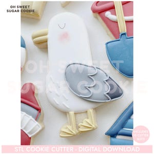 May include: A decorated sugar cookie in the shape of a seagull, with a white body, gray wing, and yellow feet. The cookie has a smiling face with pink cheeks. Other cookies in the background include a red and blue bucket and a blue shovel.