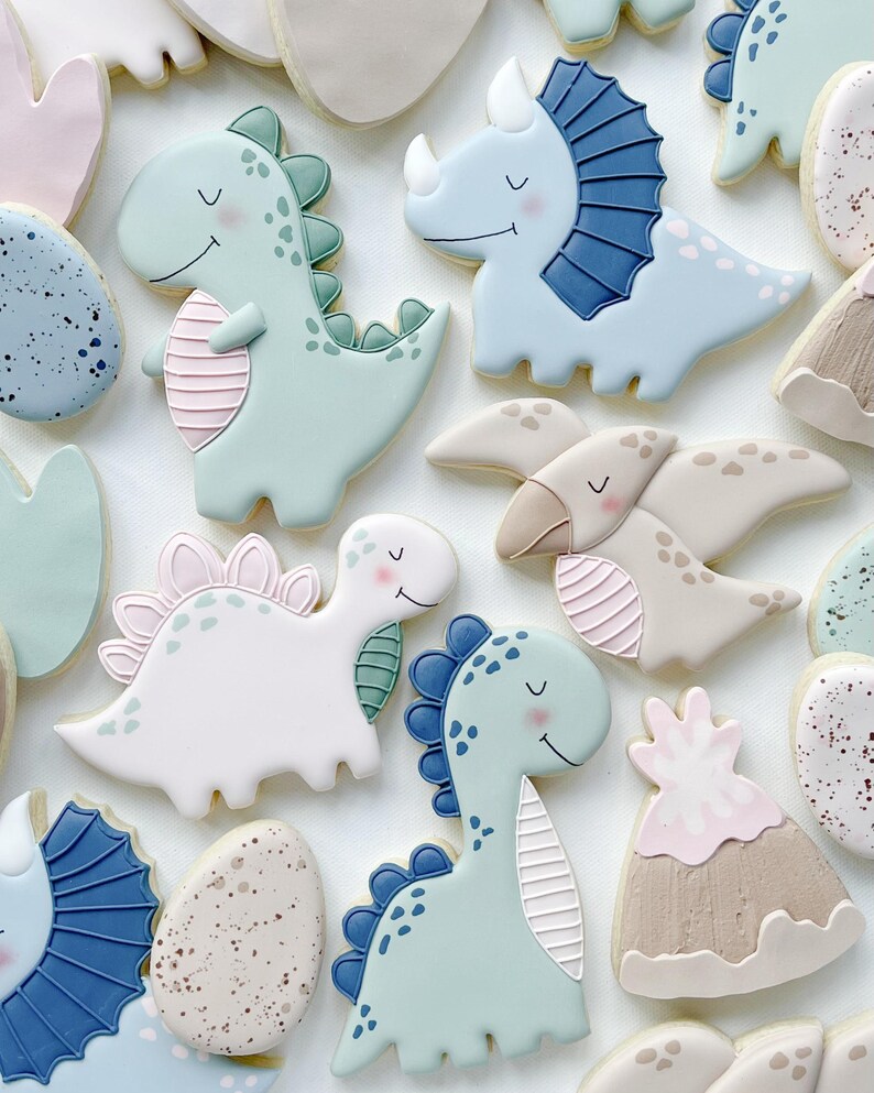May include: An assortment of dinosaur-shaped cookies in pastel colors. The cookies include a triceratops, a stegosaurus, and a T-Rex, with details like spines, horns, and stripes. Other cookies are shaped like eggs and volcanoes.
