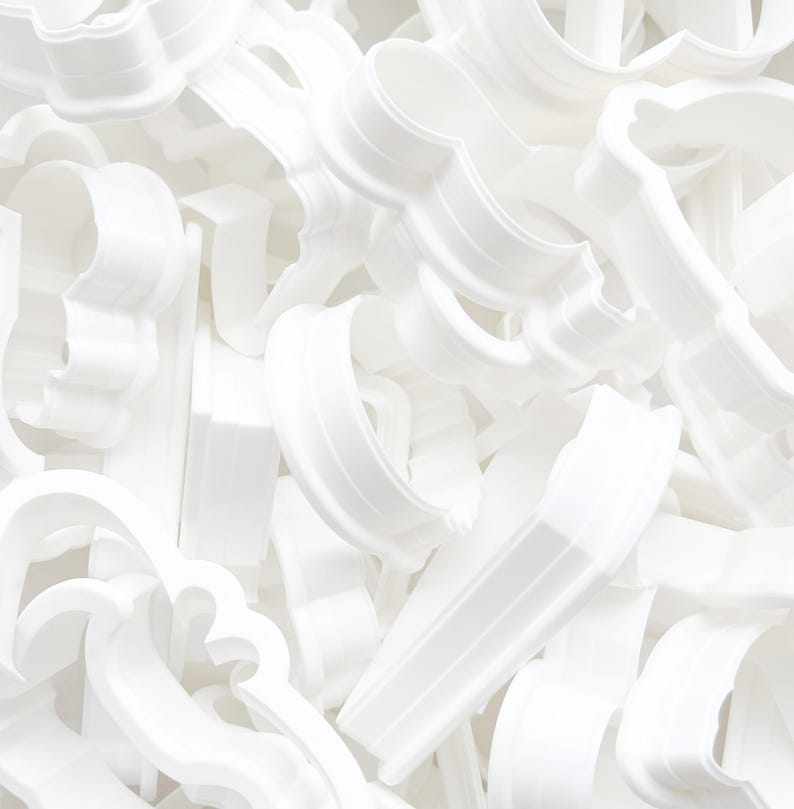 May include: A collection of white plastic cookie cutters in various shapes, including scalloped edges and curved designs. The cutters are piled together, creating a textured, monochromatic composition. Ideal for baking and crafting.