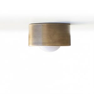 Pode incluir: Uma luminária de teto embutida com uma cúpula cilíndrica de latão. A luz tem uma lâmpada esférica branca. A luminária é montada em um teto branco. O latão tem um gradiente de cores.