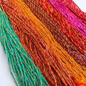 May include: A collection of multi-coloured twisted cords featuring vibrant shades of teal, orange, red, and magenta. Each cord is intricately woven with a metallic gold thread that runs throughout the length, creating a shimmering effect across the fibres.