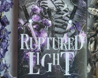 May include: A book cover for "Ruptured Light" by K. R. Richard. The cover features a dark, gothic design with a skeleton, swords, and purple floral accents. The title is in large, white, stylized letters, with the subtitle "A Destruction of Gods".