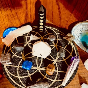 May include: A collection of crystals and gemstones arranged on a black mat with a gold geometric design. The arrangement includes a black obelisk with moon phase markings, a blue agate slice, and various crystals in shades of white, blue, and brown.