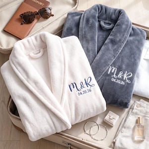 May include: Two plush robes, one cream and one dark gray, featuring embroidered text M&R and the date 04.20.26. The cream robe has navy blue embroidery, while the gray robe has white embroidery. Both robes have a shawl collar and soft texture.