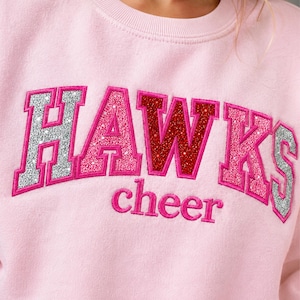May include: A light pink crewneck sweatshirt featuring the word HAWKS in large, glittery block letters with alternating silver, pink, and red colors. Below the word HAWKS, the word cheer is embroidered in smaller, solid pink lowercase letters.