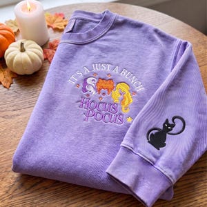 May include: A light purple crewneck sweatshirt featuring embroidered text that reads IT'S A JUST A BUNCH HOCUS POCUS above three stylized hair figures in purple, orange, and yellow. A black cat silhouette is embroidered on the right sleeve cuff.