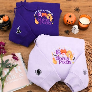 May include: A dark purple hoodie and a light lavender hoodie, both featuring embroidered text that reads It's Just A Bunch Of Hocus Pocus with illustrations of three hair silhouettes and a broom. The dark hoodie has a witch hat on the sleeve.