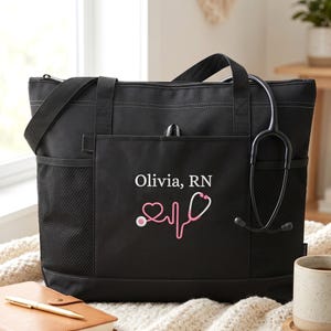May include: Black tote bag with a shoulder strap and side pockets. The bag features the name "Olivia, RN" and a pink stethoscope design. A black stethoscope hangs from the bag. A notebook and pen are in the foreground.