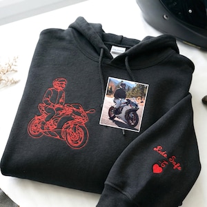 May include: A black hooded sweatshirt featuring red embroidery of a person on a motorcycle on the chest. A square photograph of a person on a motorcycle is placed on the chest. The sleeve has red embroidered text reading Ride Safe with a heart.