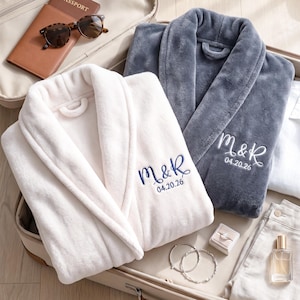 May include: Two plush bathrobes, one white and one gray, embroidered with initials and a date. The robes are folded in an open suitcase with a passport, sunglasses, jewelry, and perfume.