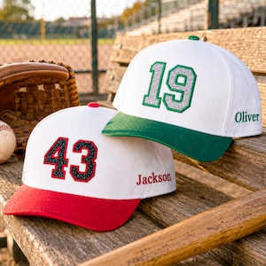 May include: Two white baseball caps with coloured brims and embroidered numbers. One cap has a red brim and the black glitter number 43 with the name Jackson. The other has a green brim and the silver glitter number 19 with the name Oliver.