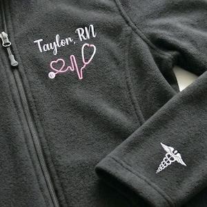 May include: A dark grey fleece jacket with a full zip. The jacket has the name "Taylor, RN" embroidered in white script, and a pink stethoscope design. A medical caduceus is embroidered on the sleeve.