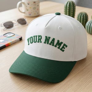May include: A white baseball cap with a green brim and a green button on top. The words "YOUR NAME" are embroidered in green on the front of the cap. The cap is sitting on a wooden surface.